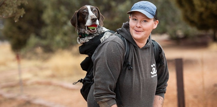 Staff member wearing hat and carrying a dog in a backpack