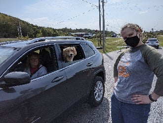 Woman in gray sweatshirt standing next to car with white dog in back seat