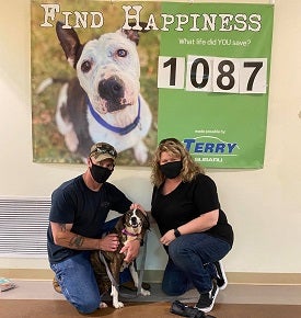 Couple wearing black masks kneeling with dog between them in front of shelter banner