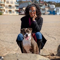 Woman wearing sunglasses crouching behind brown pit bull type dog on the beach