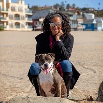 Woman in sunglasses and dark coat squatting with brown and white dog on beach