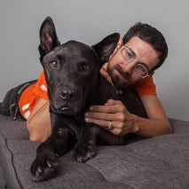 Man in orange shirt lying on gray dog bed with black dog