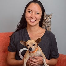 Woman in sitting in orange chair holding tan and white dog and with a kitten on her shoulder