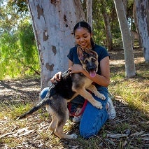 Woman in dark shirt and jeans squatting in front of a tree and hugging a German shepherd puppy
