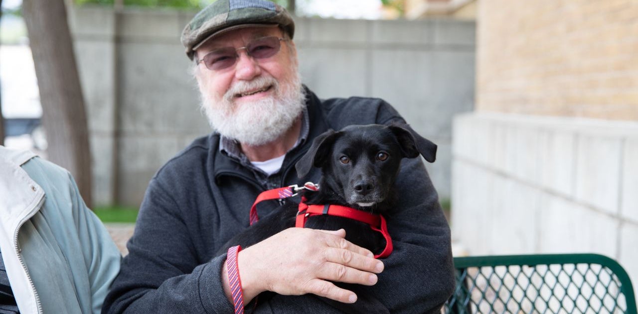 A man with a hat, glasses and white beard sits on a bench while holding a newly adopted black dog wearing a red harness and leash