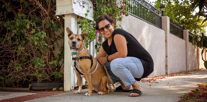 Lady in black shirt and jeans squatting next to tan dog in front of white fence