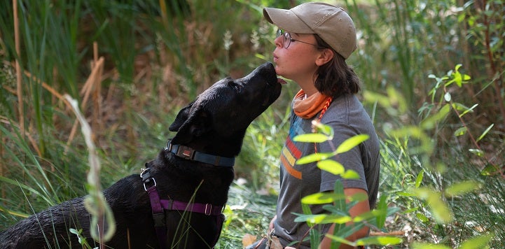 Woman in gray shirt and baseball cap having face sniffed by black dog in a field