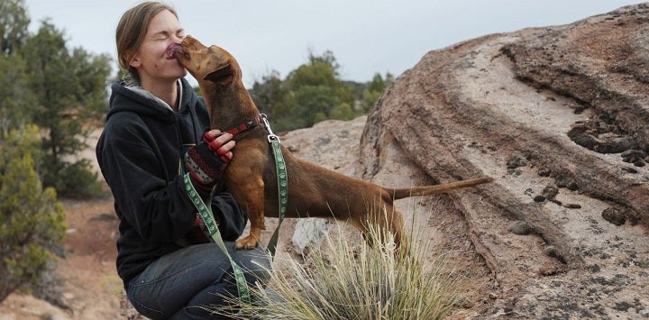 Blond woman crouching down with brown dog sniffing face with mountains in background