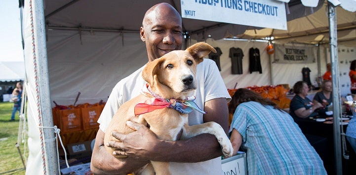 Smiling man in gray shirt holding tan dog wearing orange bandana in front of adoption booth